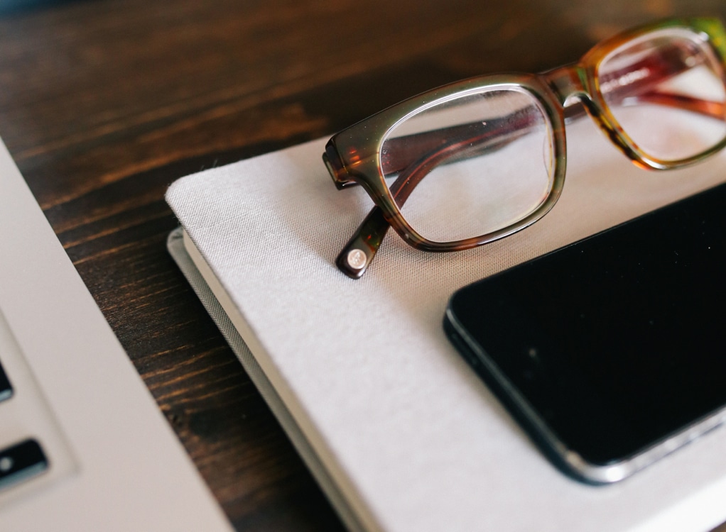 image of glasses and an iPhone sitting on a grey notebook atop a wooden desk