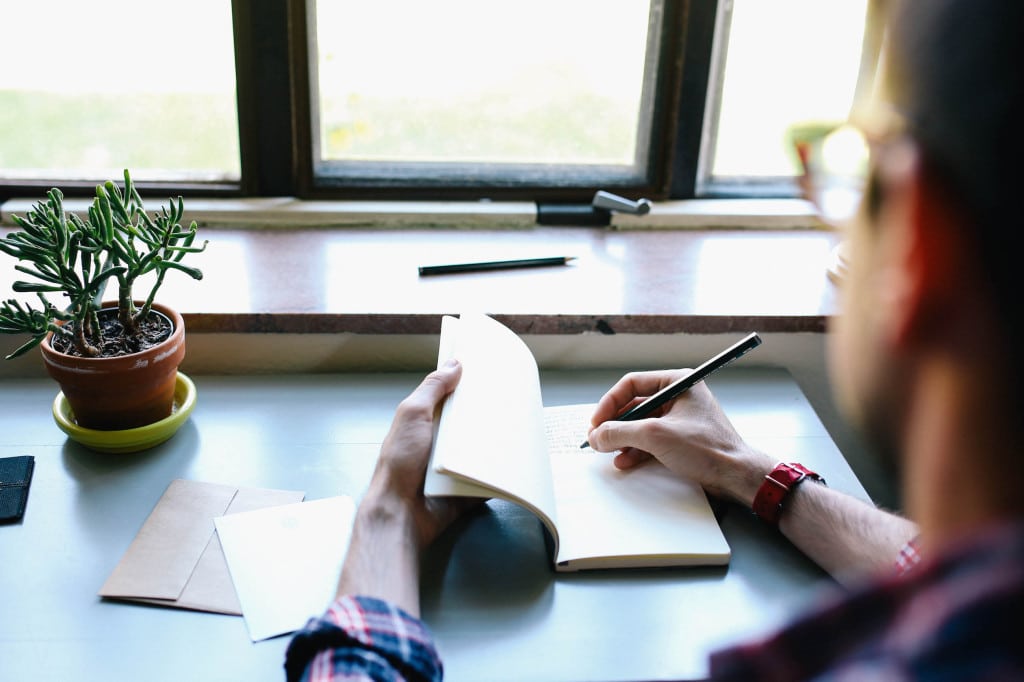 a man jots down notes in a small notebook at a desk in front of a window. to the left of a desk sits a small plant