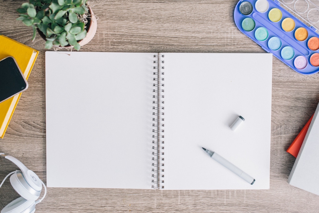 an open, blank spiral notebook with an open marker, watercolors, a plant, a book, a cell phone, and headphones also on the desk