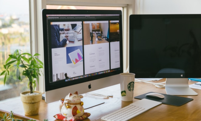 Two monitors on a desk with a laptop and a plant