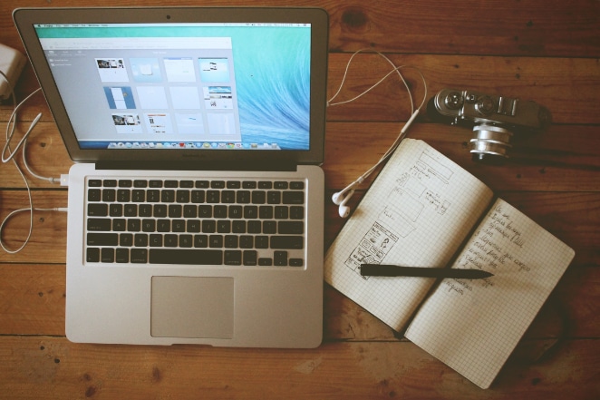 a laptop with headphones plugged in sits open on a wooden desk next to an open notebook, pen, and camera