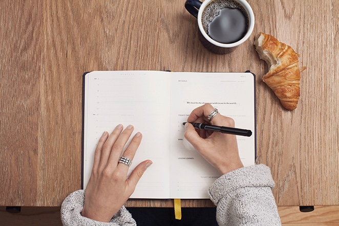 layout by flywheel recurring revenue freelance business woman writing in planner with coffee and snack on wooden table