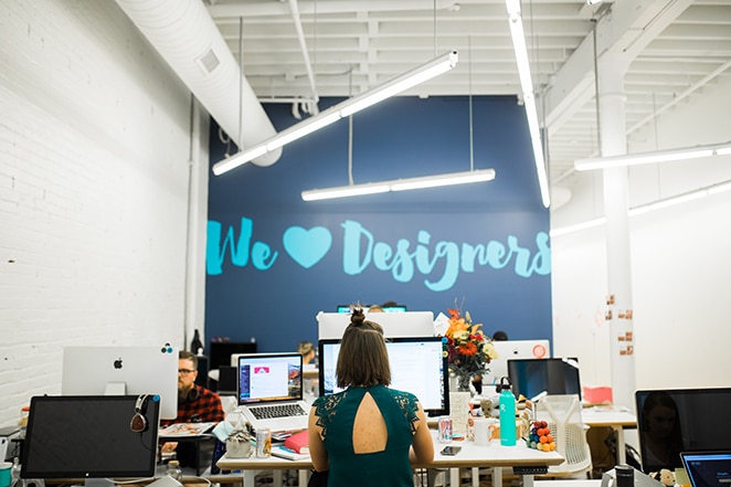 woman at desk in front of big blue wall with we heart designers painted on it