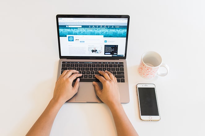 person typing linkedin webpage on computer on white desk with coffee cup and iphone nearby