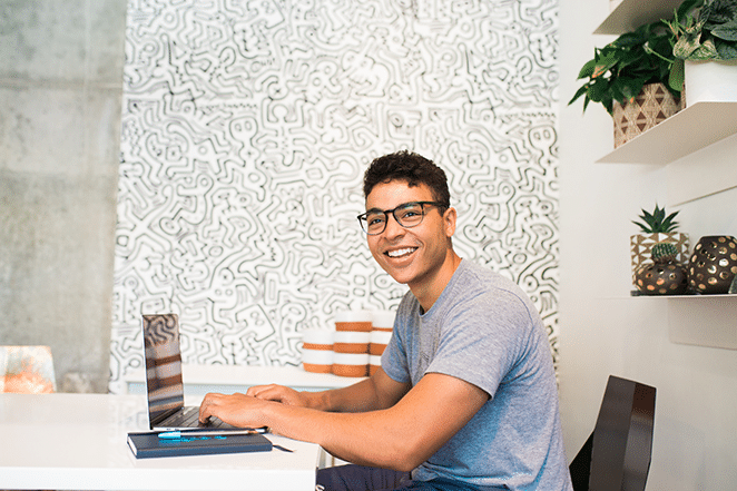 a man smiles at the camera with hands poised to type on his laptop. He sits in front of an elaborately decorated wall